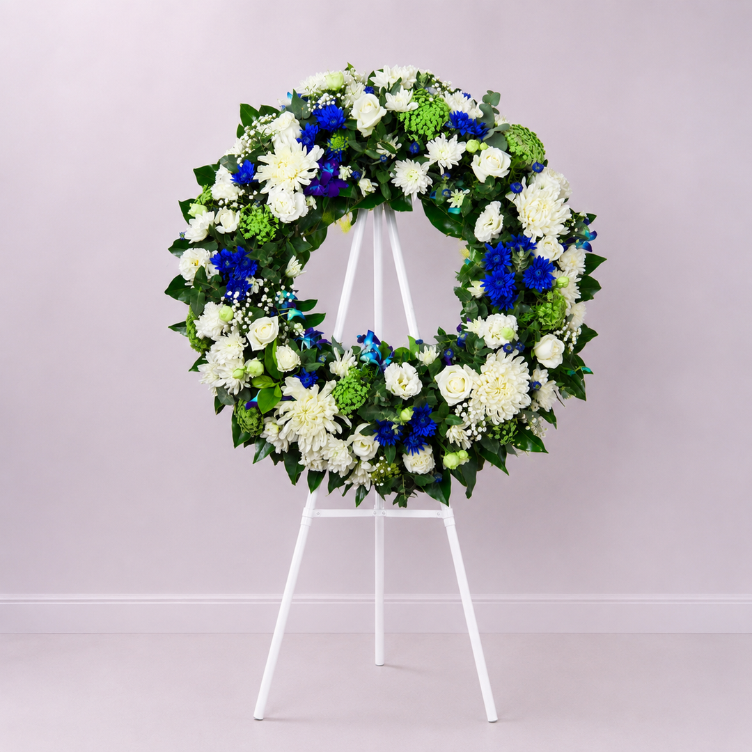 Blue Serenity funeral wreath featuring white roses, blue seasonal flowers, chrysanthemums and fresh greenery arranged in a classic circular tribute.