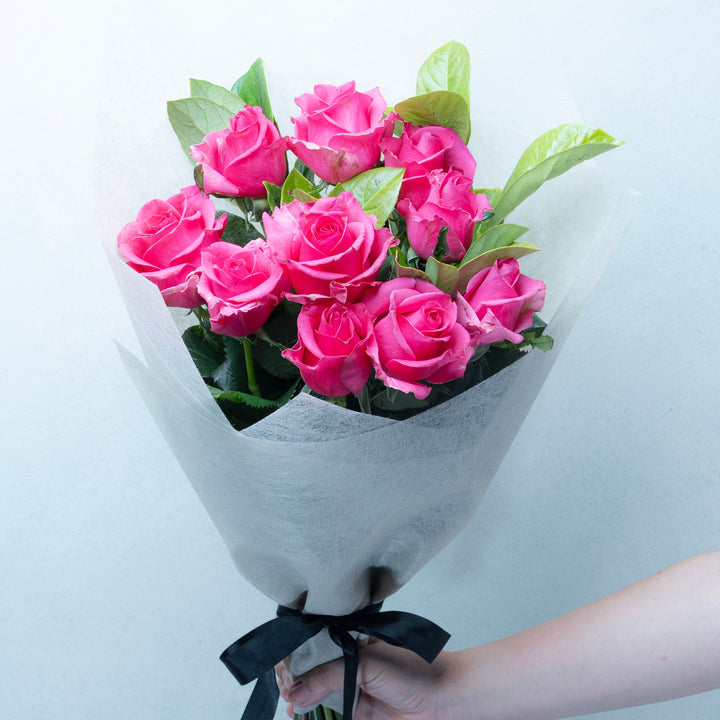 Bouquet of pink roses wrapped in white paper with a black ribbon, held by a hand against a light gray background.