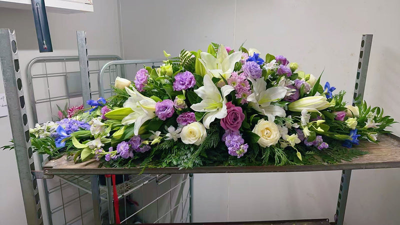 A large floral arrangement for a casket, featuring white lilies, purple roses, lavender sprigs, blue flowers, and green foliage, displayed on a metal cart in a storage room.