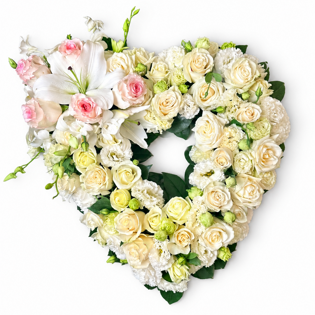 A stunning heart-shaped funeral wreath featuring an arrangement of white roses, blush-pink-tipped roses, large white lilies, white lisianthus, and greenery, presented against a clean white background. This elegant floral tribute signifies pure love and grace.